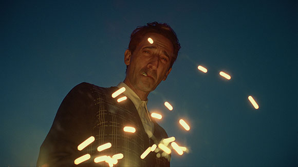 A man in a dark patterned suit looks upward amidst floating, glowing white streaks against a dark blue sky.
