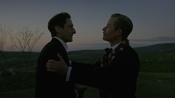 Two men in dark suits facing each other against a twilight landscape.