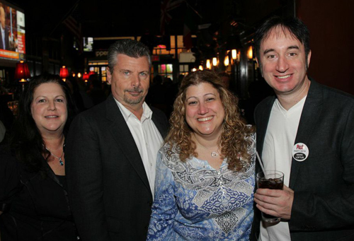 Four smiling adults posing together in a dark indoor venue with warm lighting in the background.