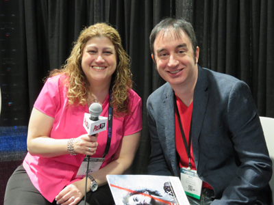 A smiling woman in a pink shirt holding a microphone sits next to a smiling man in a blue blazer.