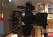 A large, professional black video camera mounted on a tripod in a dimly lit room.