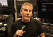 A man in a black t-shirt holds a microphone in front of studio monitors.