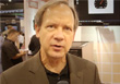 A man with light hair wearing a brown suit jacket and black shirt looks toward the camera.