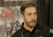 A man with light brown hair and a beard speaks into a microphone in front of a poster.