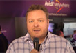 A man in a plaid shirt speaks into a black microphone against a purple background.