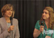 Two women holding microphones and speaking in front of a dark curtain.