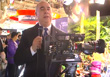 A man in a dark suit and blue tie speaks into a microphone in front of a large video camera.