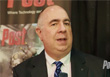 A bald man in a dark suit and blue patterned tie speaking.
