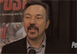 A man with dark hair and a goatee speaking into a microphone against a dark background.