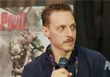 A man with short brown hair and a mustache holds a microphone while looking to the side.
