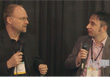 Two men in blazers hold microphones and face each other while talking against a dark curtain.