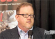 A man with short reddish hair and glasses wearing a suit jacket and light blue shirt.