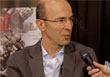 A man with glasses and a grey suit jacket speaks into a microphone held by an unseen person.