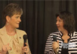 Two women sit facing each other, holding microphones against a dark curtain background.