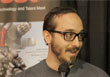 A man with dark hair, glasses, and a beard speaks into a microphone against a blurred background.
