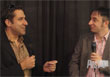 Two men in blazers smiling and talking to each other against a dark background.