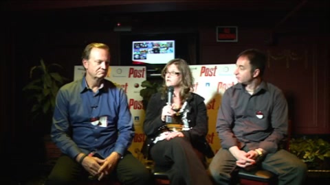 A man in blue, a woman with a microphone, and a man in grey sit on a stage in front of a Post backdrop.