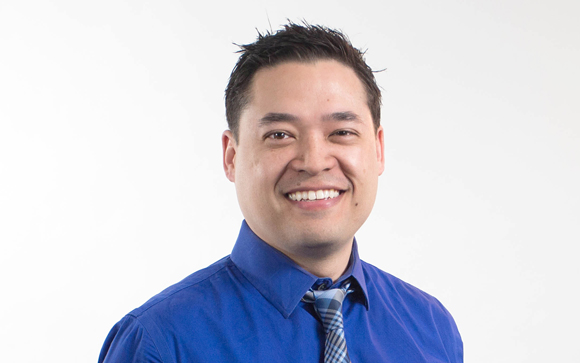 A smiling man with short dark hair wearing a blue button-down shirt and a patterned blue tie.