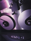 Close-up view of two large metal reels on a reel-to-reel tape recorder under purple lighting.