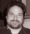 A close-up, sepia-toned portrait of a smiling man with dark wavy hair and a beard.