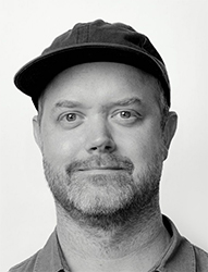 Black and white headshot of a man with a beard wearing a dark baseball cap.