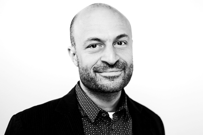 Black and white headshot of a bald man with a beard and a patterned shirt.