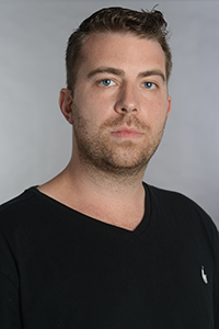 A man with short brown hair and facial stubble wearing a black V-neck shirt.