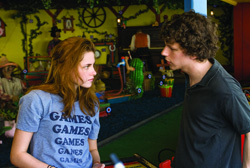 A young woman in a blue t-shirt and a young man in a dark polo shirt face each other in a colorful arcade.