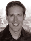 A smiling man with short hair wearing a dark striped collared shirt against a blurred cityscape background.