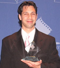 A man in a dark suit and pink tie smiling while holding a glass award trophy.