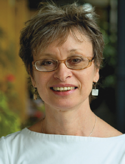 A smiling woman with short brown hair and glasses wearing a white top and dangling earrings.