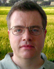 A man with short brown hair and glasses looking at the camera against a background of a yellow field.