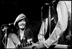 Black and white photo of a smiling man in a flat cap performing music on stage next to a guitarist.