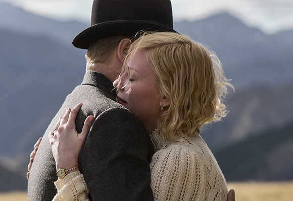 A man wearing a black top hat and grey coat hugs a blonde woman in a white lace dress against a mountain backdrop.