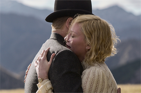A man in a top hat and grey coat hugs a blonde woman in a white lace dress against a mountain backdrop.