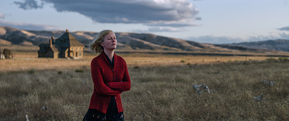 A blonde woman in a red cardigan stands in a dry field with her eyes closed against a backdrop of hills.