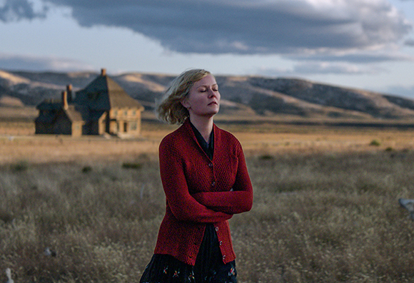 A blonde woman in a red cardigan stands with eyes closed in a grassy field before a large house.