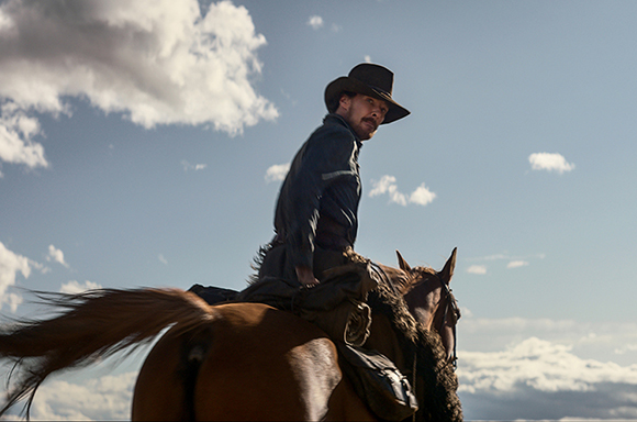 A man wearing a dark cowboy hat and blue shirt rides a brown horse against a cloudy sky.