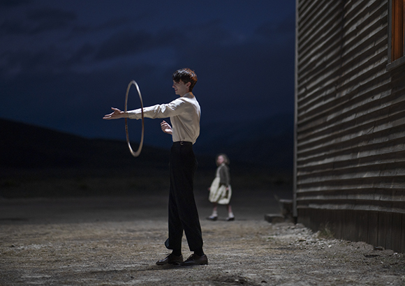 A person in a white shirt and dark trousers spins a silver hoop in a dark field at night.
