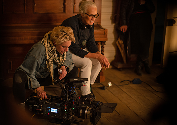 Two women crouch over a professional film camera on a wooden floor in a dimly lit room.
