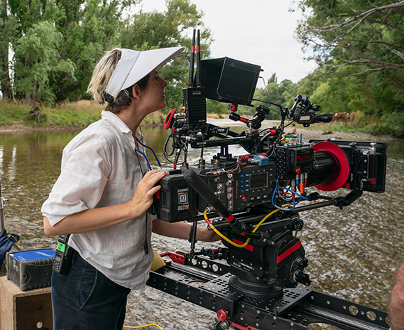 A person in a white shirt and visor looks into a large professional cinema camera mounted on a track by a river.