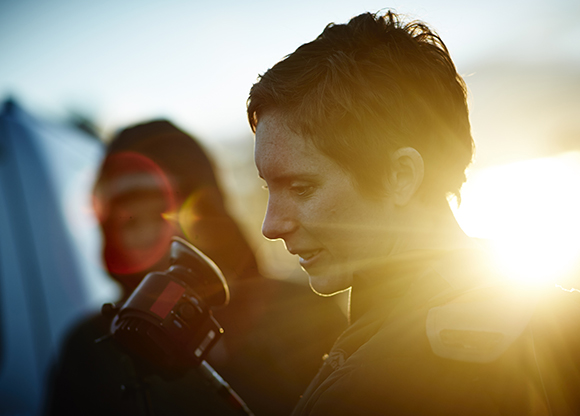 A person with short hair looks down at a camera held in front of them during a bright sunset.