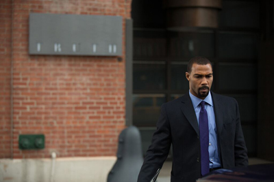 A man in a dark suit and blue tie stands outdoors in front of a brick building with a sign that reads 'TRUTH'.
