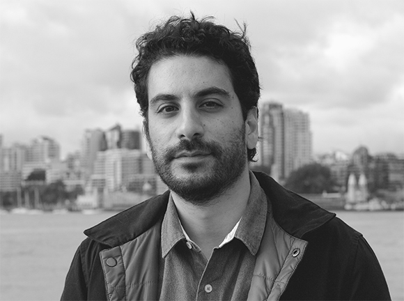 Black and white portrait of a man with dark curly hair and a beard, with a blurred city skyline in the background.