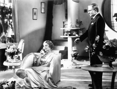 A man in a dark suit holds out a small doll toward a woman sitting in a large armchair.