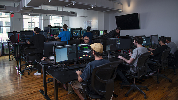 Several people sit at desks with multiple monitors in a bright, open-plan office with wooden floors.