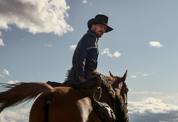 A man wearing a dark hat and jacket rides a brown horse against a cloudy blue sky.