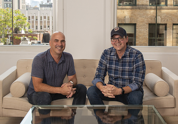 Two men smiling while sitting on a beige sofa in a bright room with large windows.