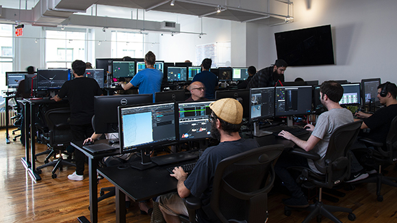 Several people sit at desks with multiple computer monitors in a bright, open-plan office space.
