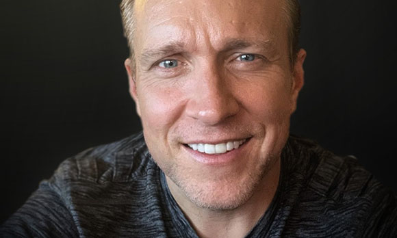 A close-up portrait of a smiling man with light eyes and short blonde hair against a dark background.